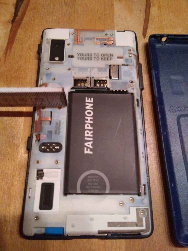 Die Rückseite eines Fairphone 2 mit abgenommenem Deckel. Der Akku ist zu sehen. Ausserdem ein Zollstock.