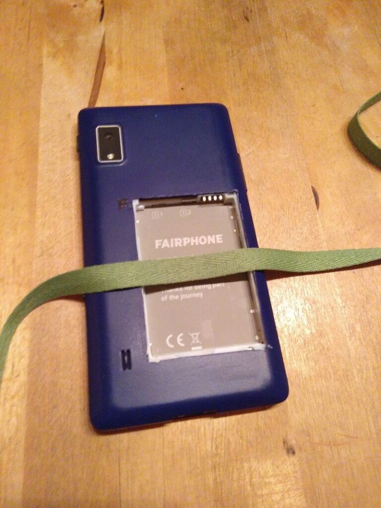 Die Abdeckung wird zurück auf das Fairphone gelegt. Die Abdeckung hat nun hinten ein Loch in Grösse des Akkus. Ein grünes flaches Band, ca. 20cm lang und 1cm breit, wird von links nach rechts über das Loch hinten auf das Smartphone gelegt.