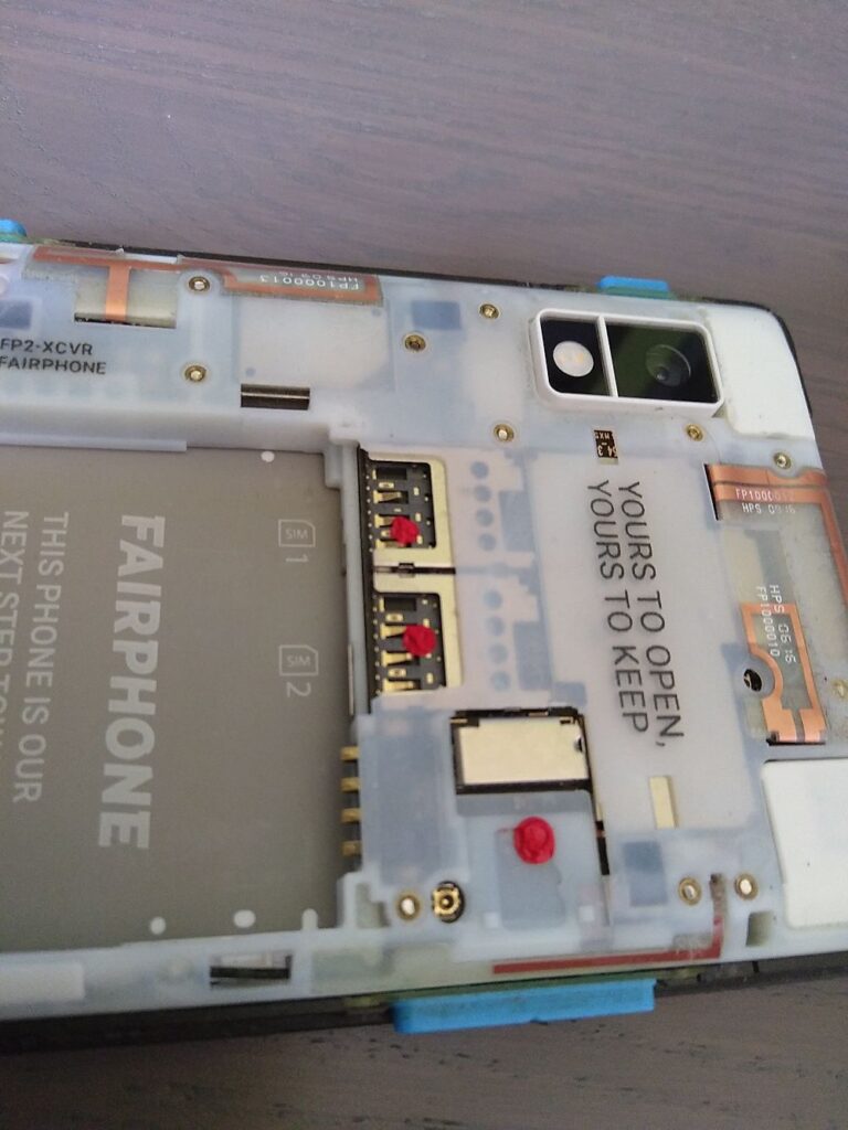 Zu sehen ist die Rückseite eines geöffneten Fairphone2 in Nahaufnahme ohne Akku. Dort, wo die Simkarten bzw. die Speicherkarte eingelegt werden wurde jeweils ein roter tropfen Siegellack angebracht. Der Lack ist hart und fühlt sich kantig an.